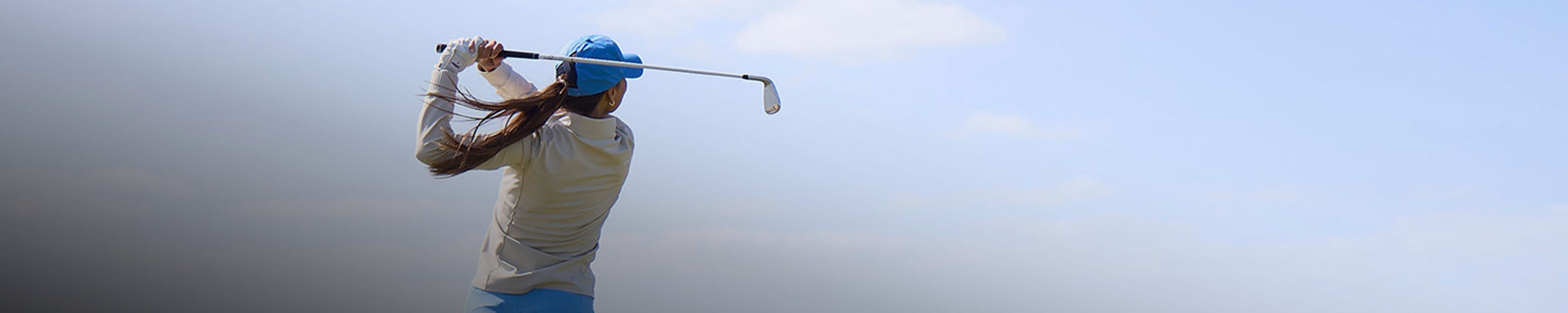 A woman wearing a KJUS midlayer swings a golf club in front of a clear blue sky.